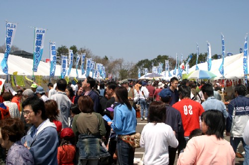 晴れた青空の下、多数の白いテントと「潮騒まつり」と書かれた青い旗が並ぶ大規模な野外会場で、様々な年齢層の非常に多くの人々が活発に交流し賑わっているイベント会場の様子の写真