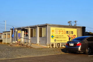 仮設の建物の外壁に「法テラス南三陸」と書かれた黄色い看板が掲示されており、玄関前には手すりとスロープが設置されている法テラス南三陸事務所の外観写真