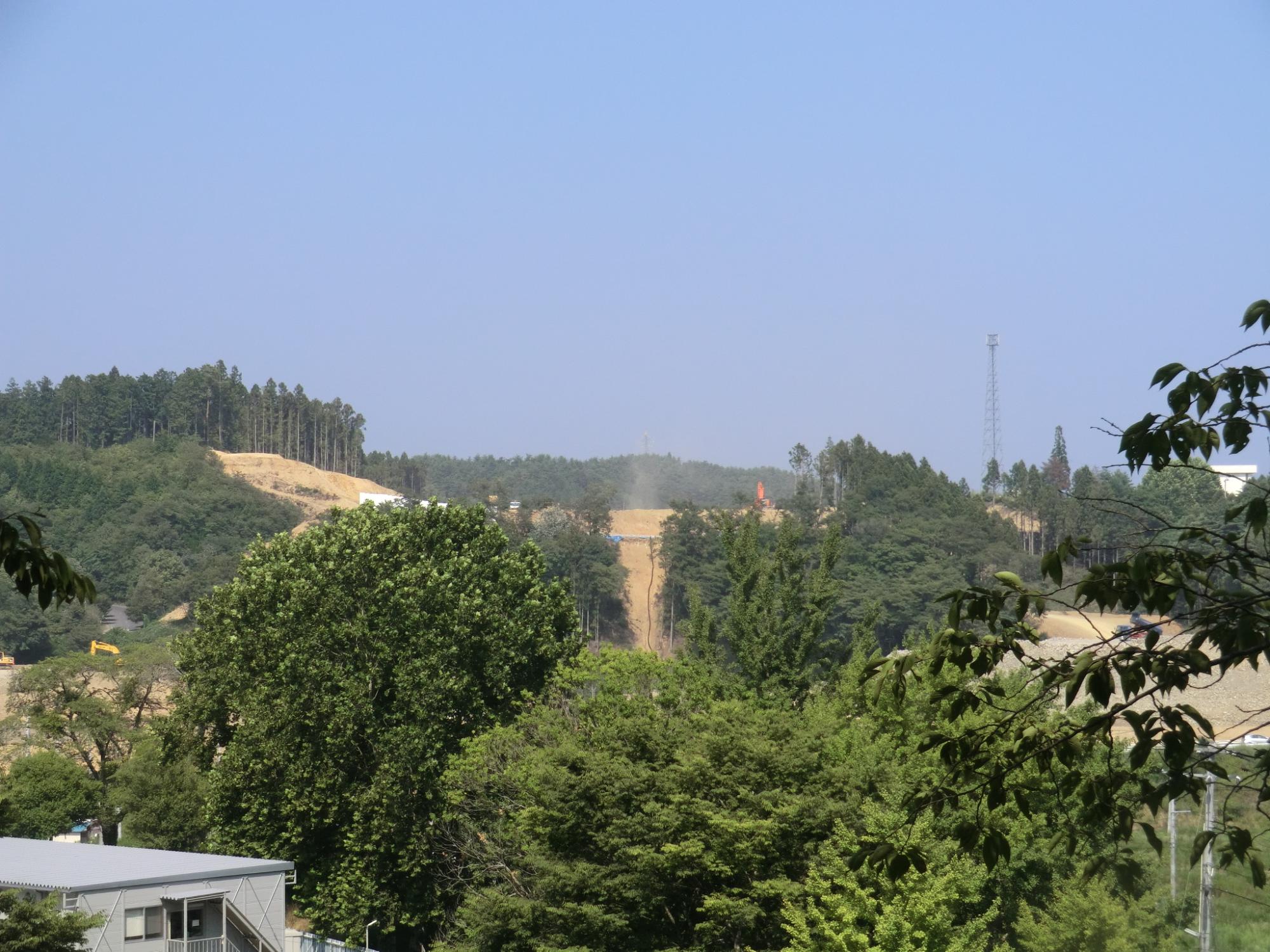 青々とした木々の葉の奥に段状に盛られた土が見え、重機が建設予定地の斜面を整地している様子が映っている写真