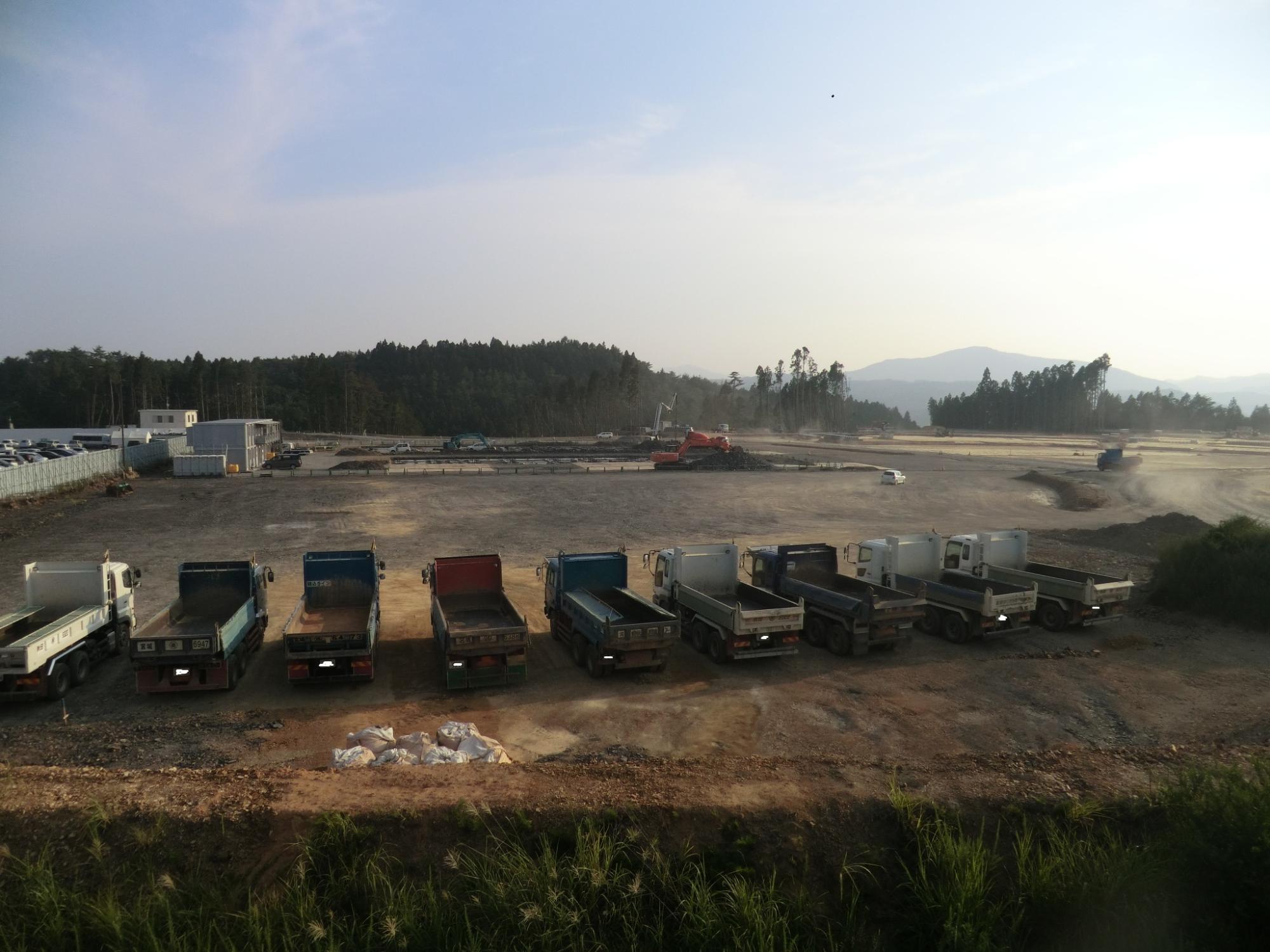 広大な造成地の手前に整然と並ぶ複数台のダンプトラックが停車し、奥では重機が稼働して工事が進行している様子の写真