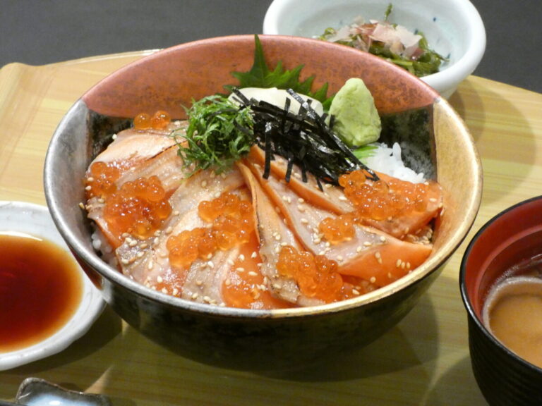 ご飯の上に厚切りのサケ刺身といくらが乗っており、上に刻み海苔・大葉・ワサビが添えられた華やかな志津川銀鮭炙り漬け丼の写真