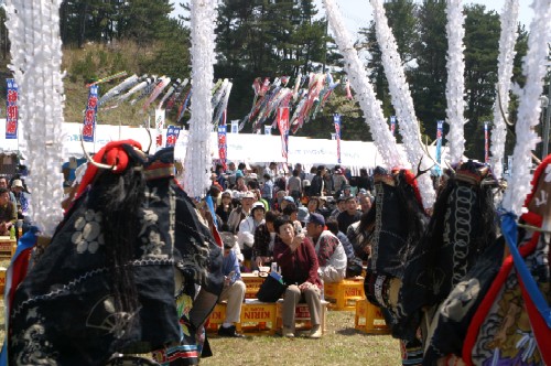 芝生の会場で角などがついた華やかな衣装の踊り手が手前に写り、その奥には大勢の観客が座って見物しているイベントの様子の写真