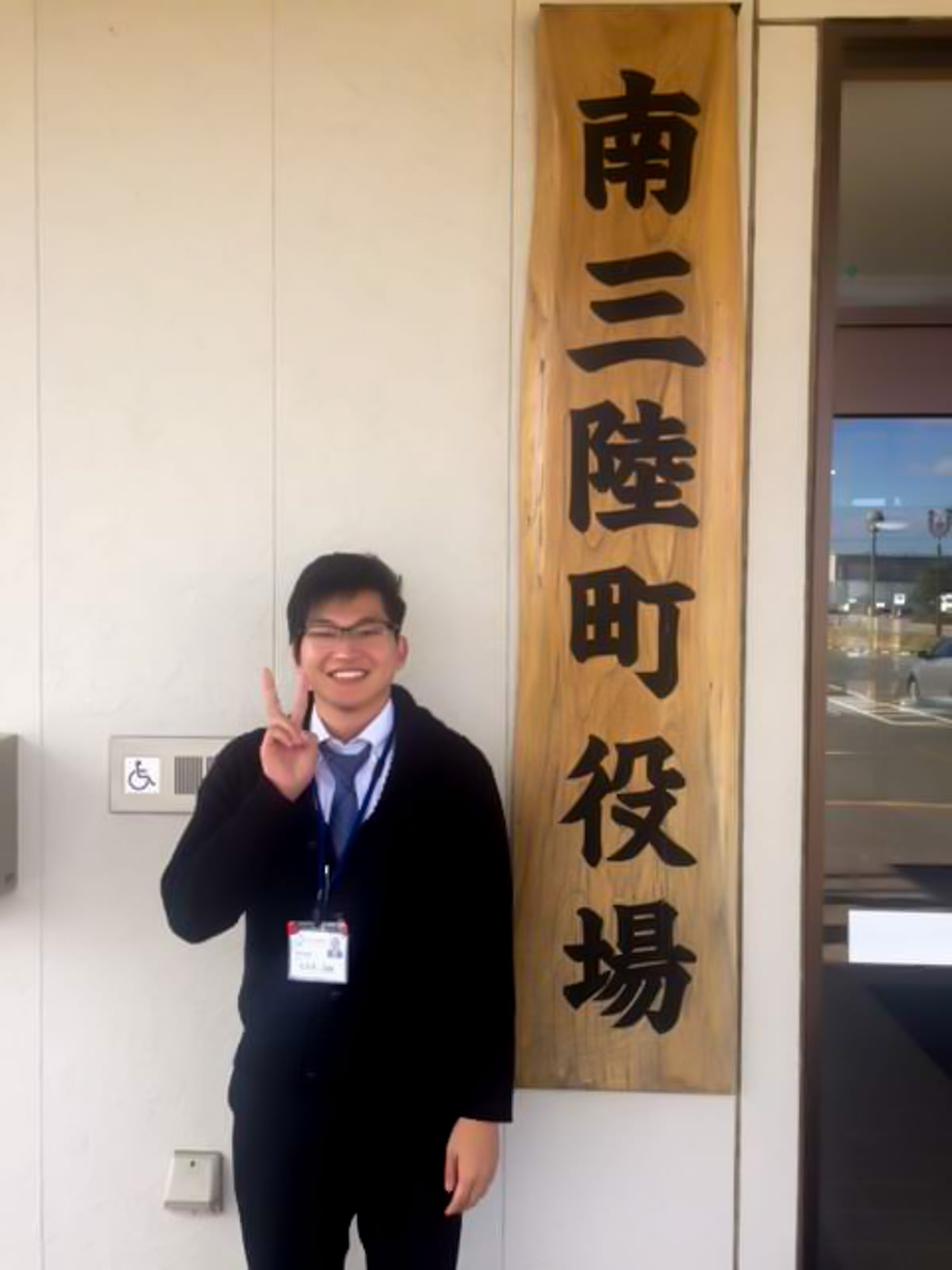 南三陸町役場と書かれた木製の大きな看板の横に立ち、ピースサインをしている佐々木さんの写真