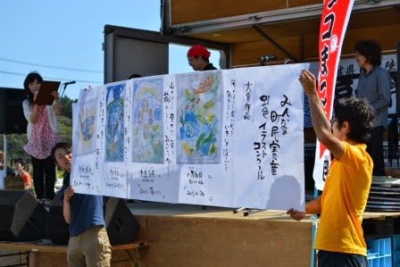 ステージの前に2人の男性が持った大きな模造紙に描かれた4枚の作品が広げられ、マイクを持った左側に立つ女性が進行を務めている授賞式の様子の写真