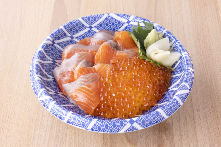 青と白の模様が入った器に、脂の乗ったオレンジ色のサケの刺身と透明感のあるいくらが半々に盛られ、横にワサビ・大葉・スライスしたガリが添えられた、見た目も鮮やかな昆布締め銀鮭いくら丼の写真