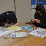 机の上で2人の生徒が紙や色見本を使って模型に必要なパーツや説明素材を作っている様子の写真