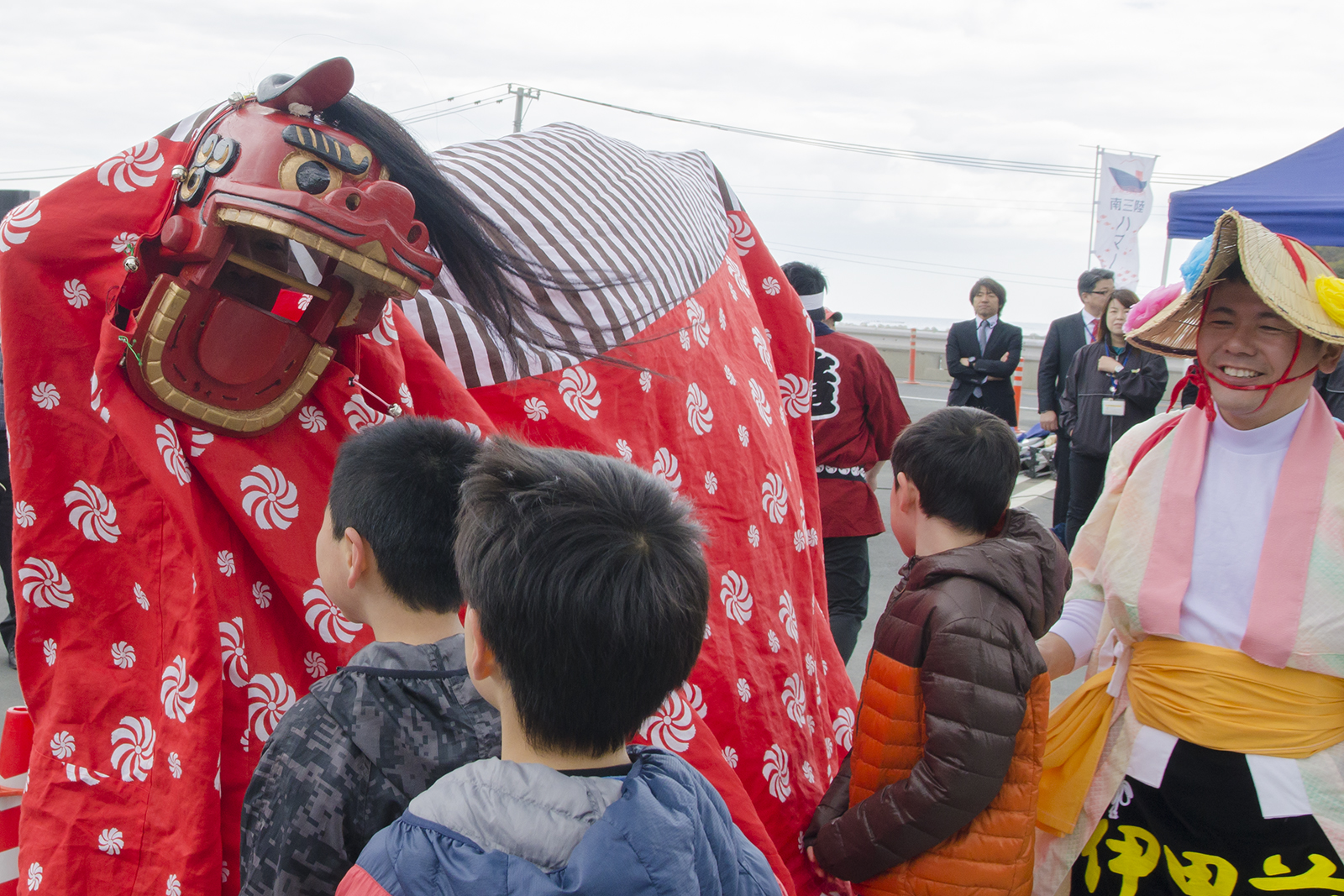 獅子舞が子どもたちの前で大きな口を開けており、笑顔の演者の男性が横に立っている写真