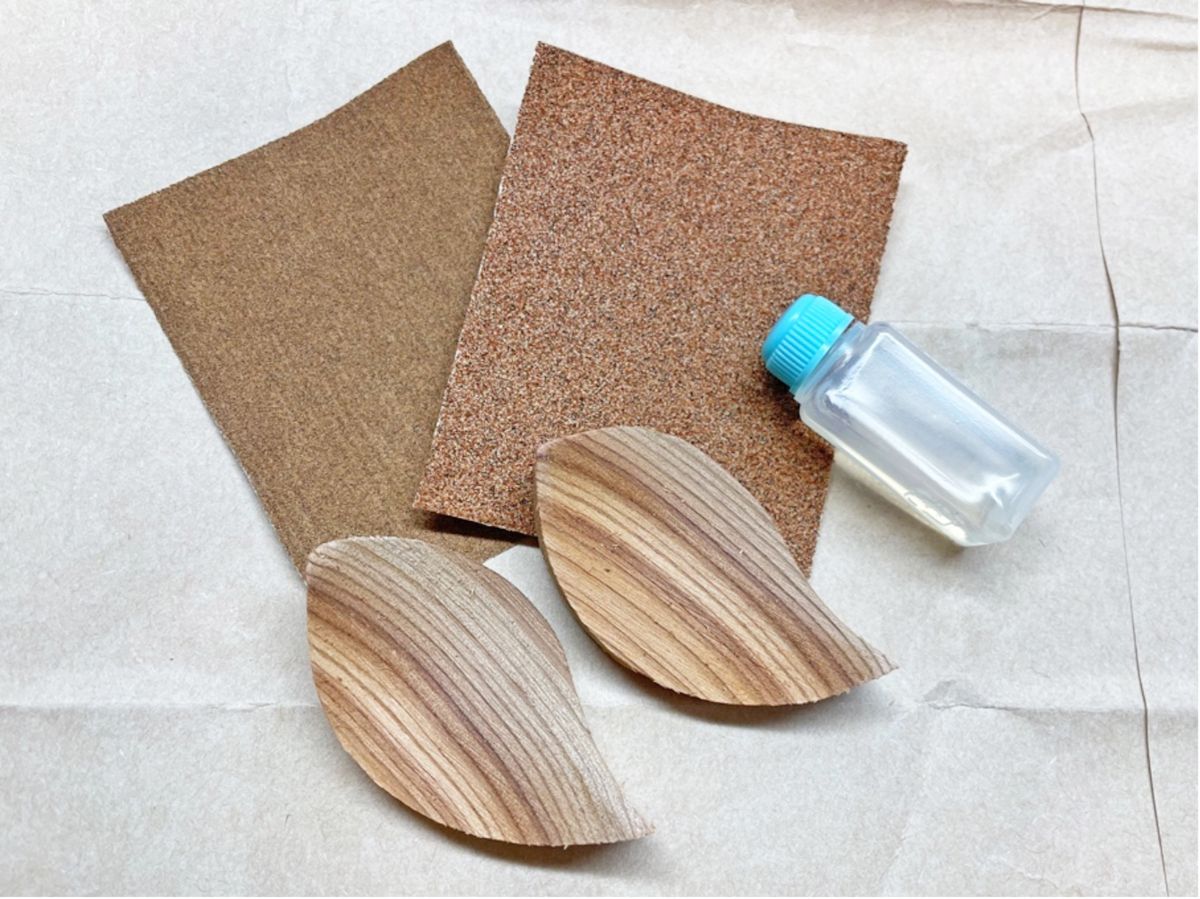 木目の美しい葉っぱ型の木材2枚と茶色の紙やすり2枚、青いキャップの小さなボトルが、白い紙の上に丁寧に並べられている写真