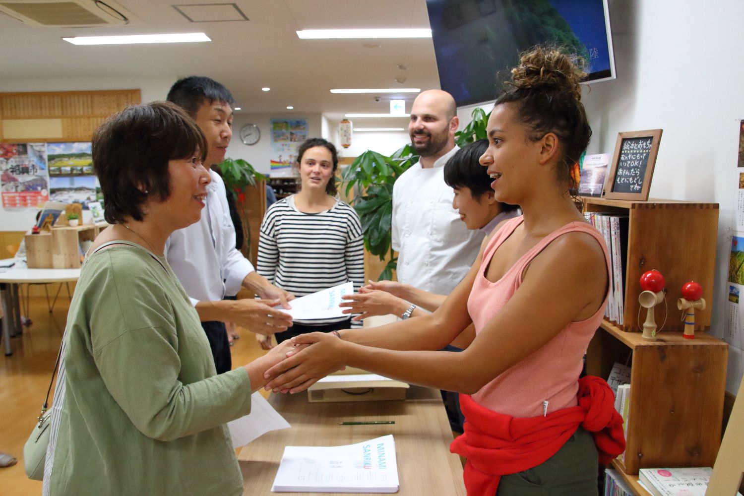 3人のシェフと1人の女性が並んで立ち、手前に立つピンク色のタンクトップ姿のシェフとお披露目会に参加した女性が握手をし、女性がシャツを着た男性に用紙を手渡している写真
