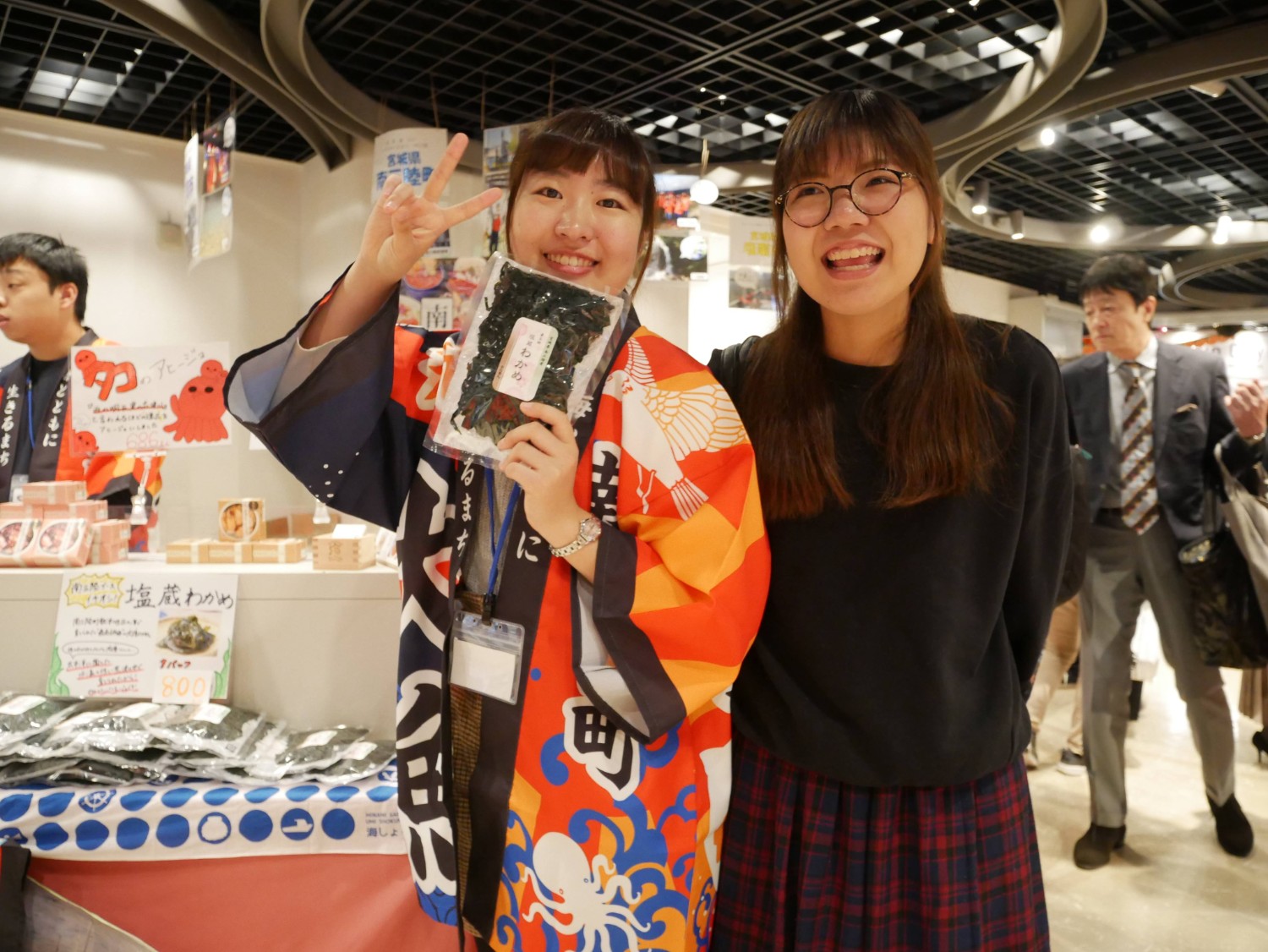 カラフルな法被を着た女性が笑顔でピースサインをしながら袋入りの「塩蔵わかめ」を手に持ち、隣で眼鏡をかけた女性が楽しそうに並んでいる店頭の様子で、後ろには「タコのアヒージョ」などの商品が並ぶ賑やかな売り場の写真
