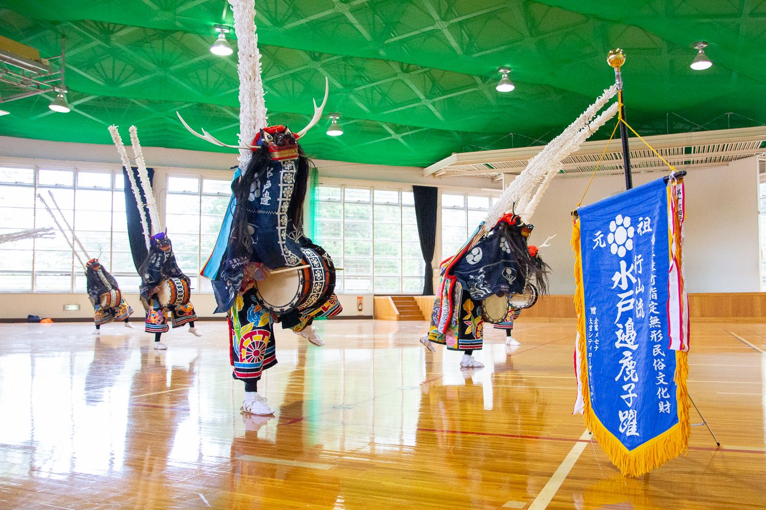 体育館の中で「水戸邊鹿子躍」と書かれた青い旗の前で、華やかな装飾と角のついた鬼の面をかぶった舞い手たちが太鼓を打ちながら迫力ある伝統舞踊を披露している写真