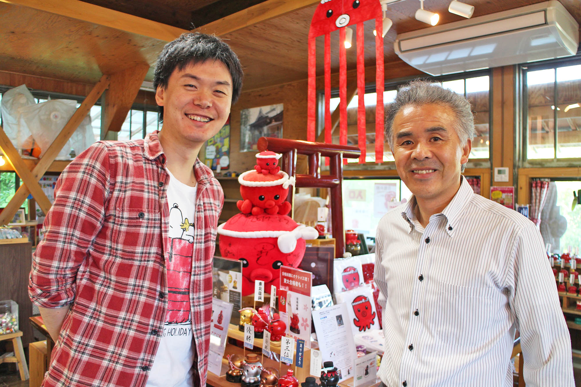 赤いタコのぬいぐるみや関連グッズがたくさん置かれた店内で、大森さんと阿部さんが笑顔で並ん写っている写真