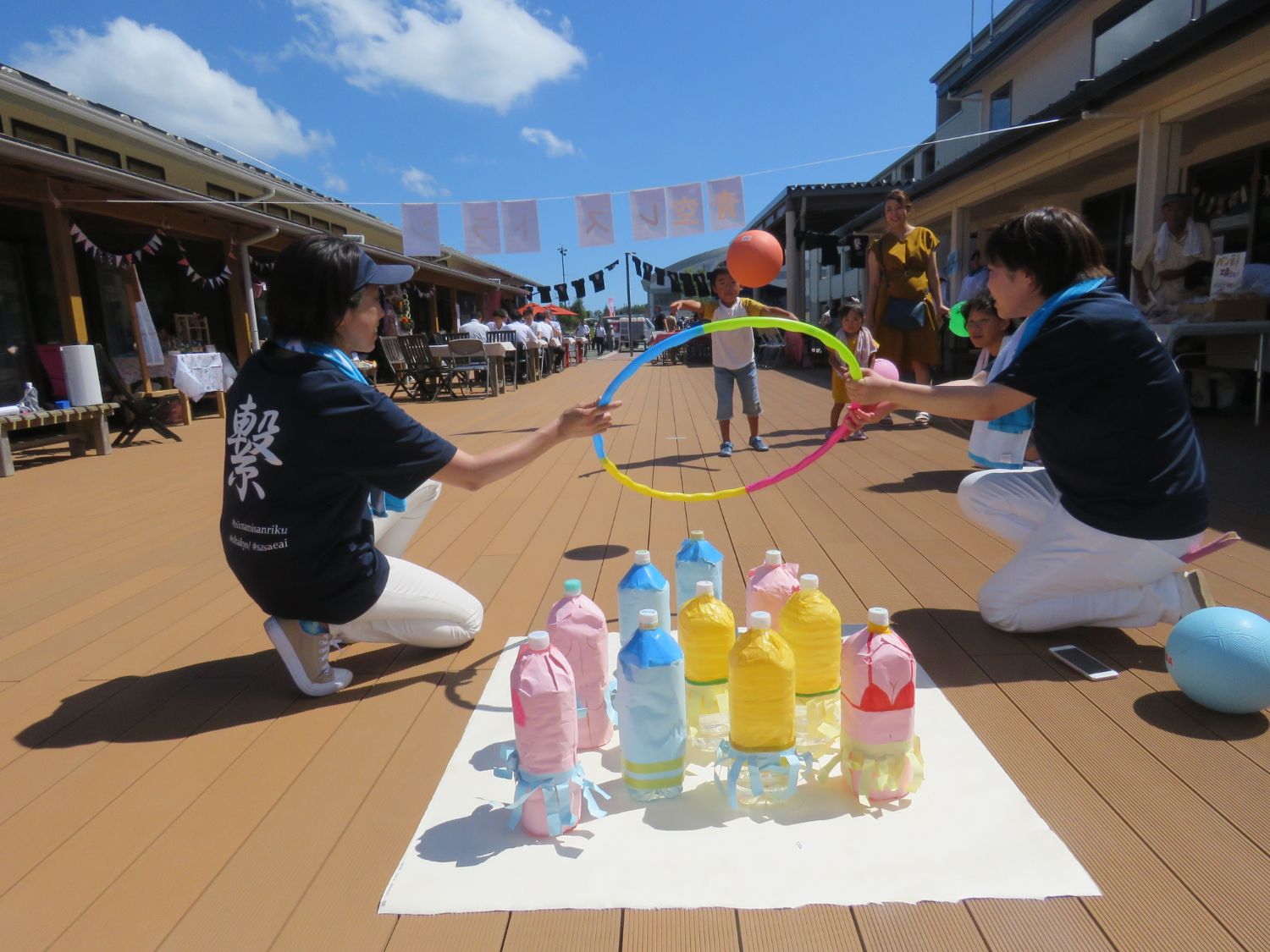 晴れ渡る青空の下、ウッドデッキの上で子どもが輪投げの輪を手に持ち、カラフルなペットボトルで作られた的に向かって投げようとしている様子を、大人たちがフラフープを使って誘導しながら見守っている屋外イベントの写真