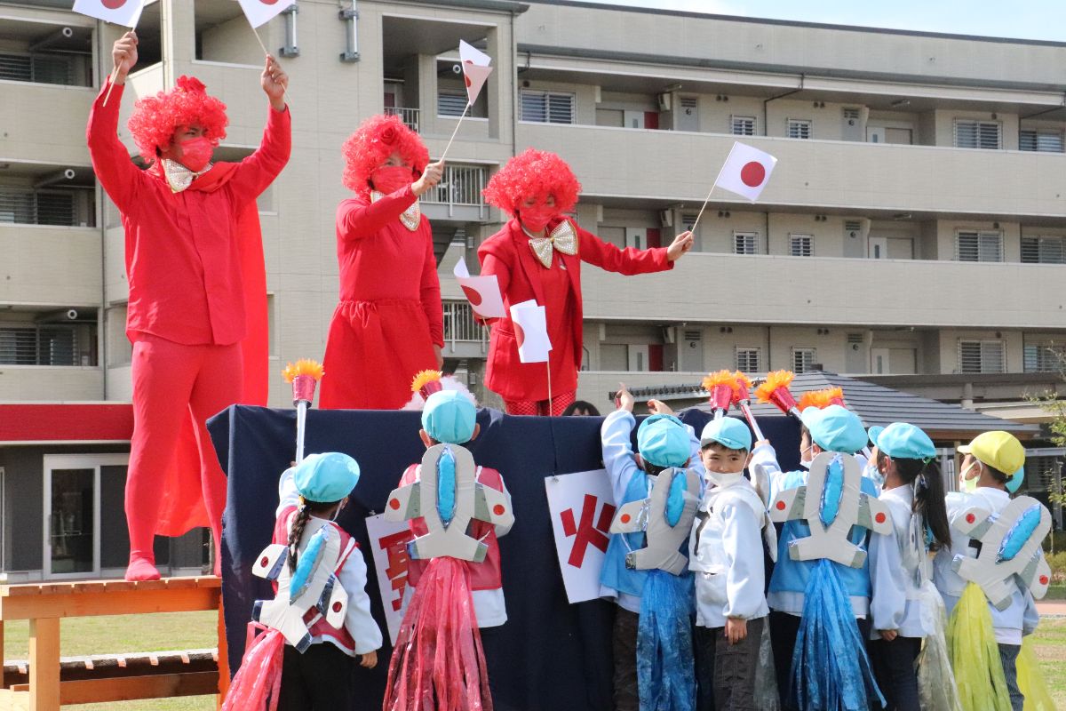 飛行士のような衣装を着た子どもたちが手作りの聖火を持ち、赤い服とかつらを身につけた3人の大人が立つステージの前に集まっている様子の写真