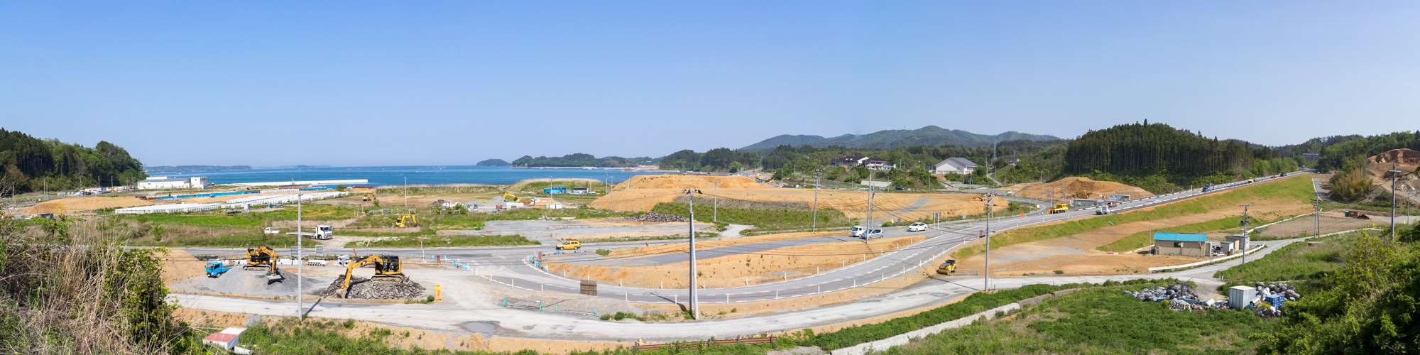 青く穏やかな海があり、海岸には白い建物や仮設のような構造物が点在している様子を定点カメラによる昼間の風景で、あちこちに黄色いショベルカーやブルドーザーなどの重機が見え広大な海岸沿いの地域を高台から見渡したパノラマ写真