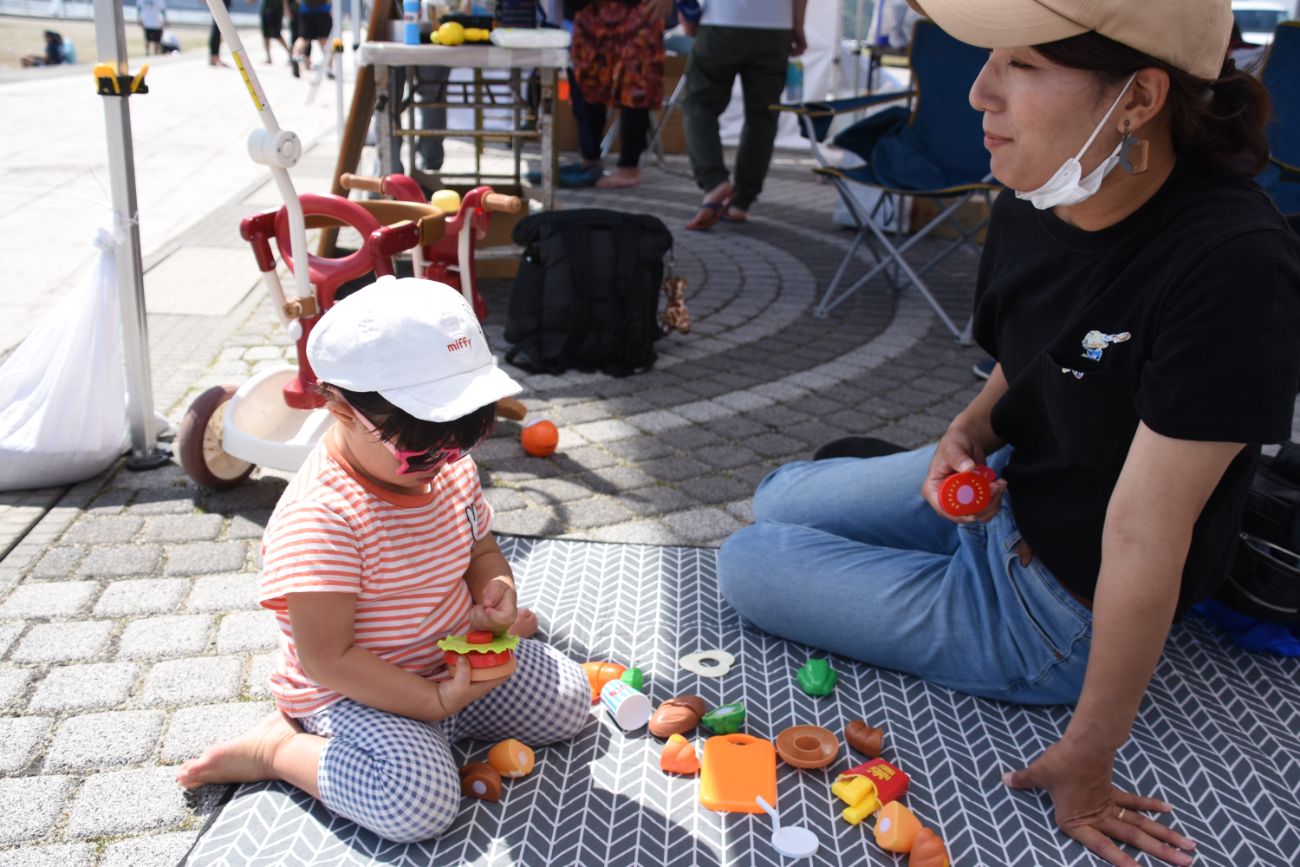 日差しの強い屋外で、子どもが白い帽子をかぶりストライプのシャツを着て、母親とともにグレーの敷物の上でおもちゃの食べ物を手に遊んでいる写真