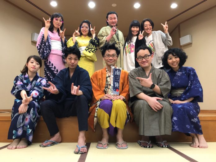 10人の男女が畳の部屋の舞台上で浴衣などの衣装を着てポーズを取り、前列中央の男性が「南三陸」と書かれた小さなのぼり旗を持って座っている写真