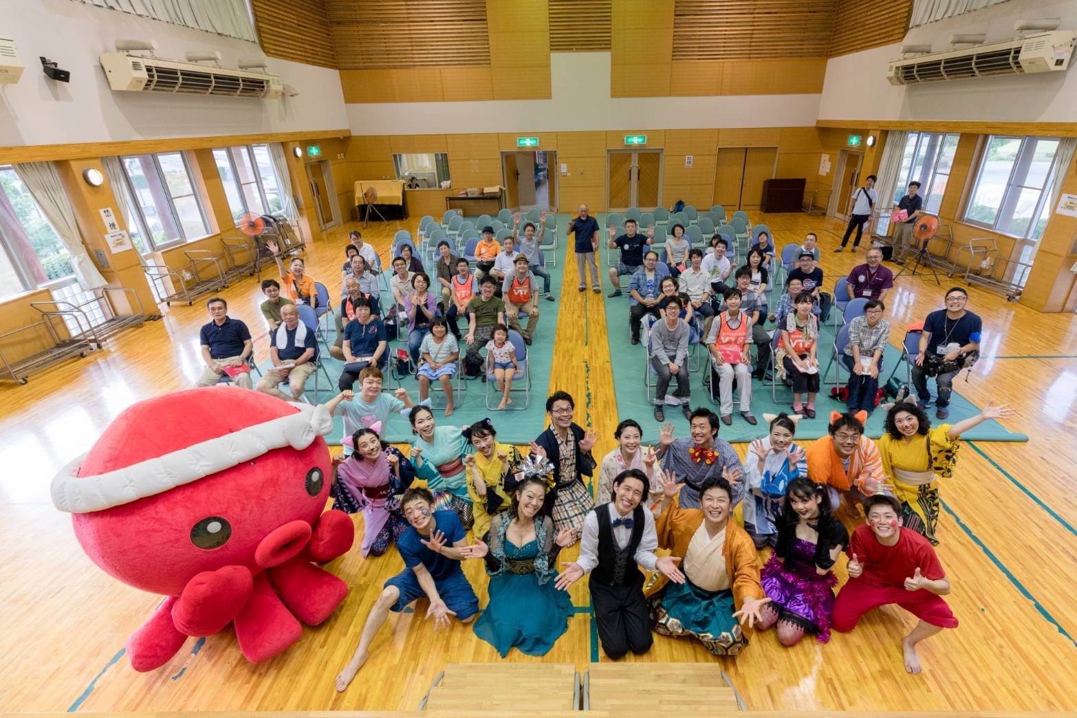 体育館で椅子に座った観客の前にオクトパス君の着ぐるみとカラフルな衣装を着た出演者が並び、全員がカメラに向かって手を振っている写真