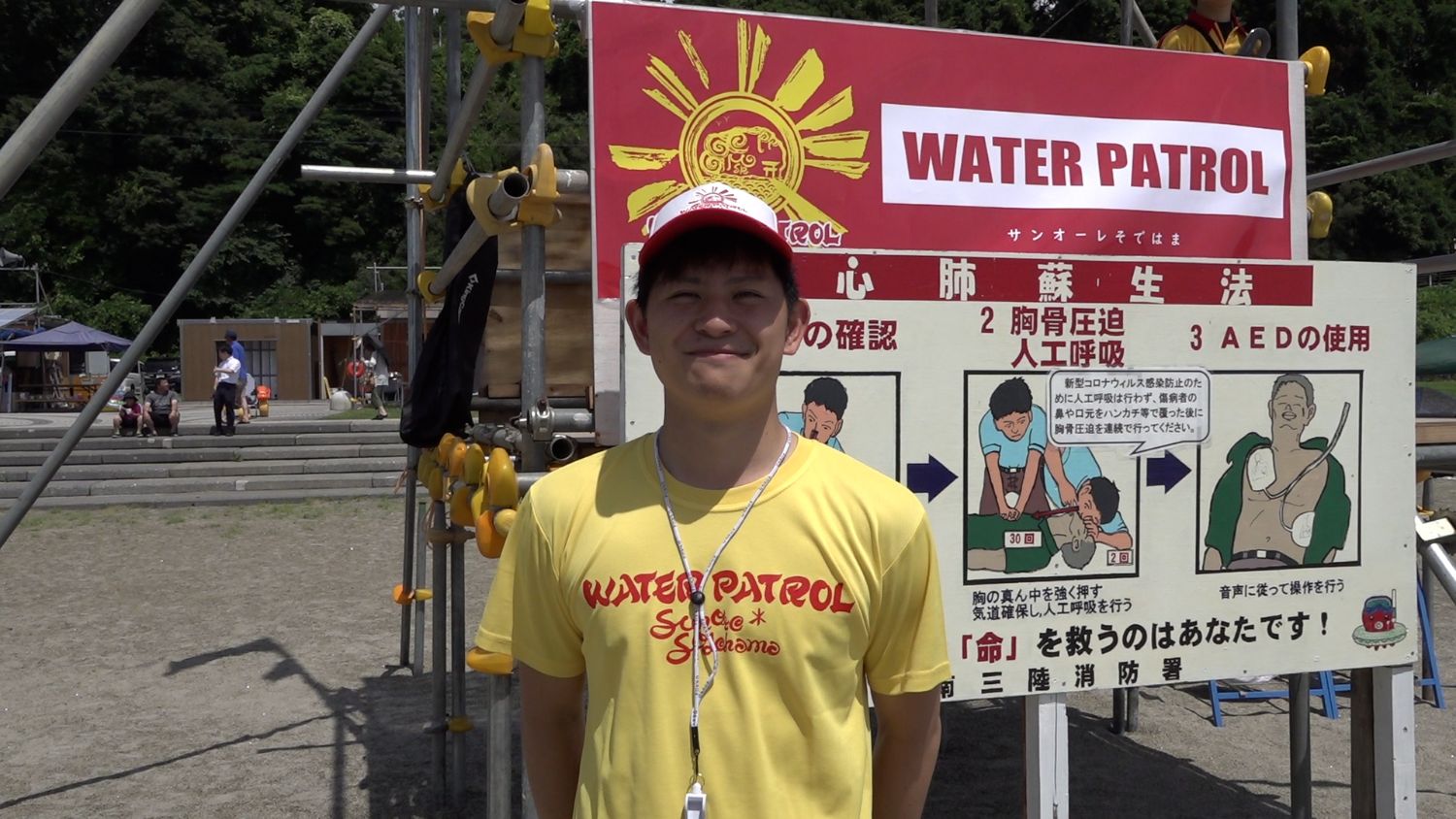 黄色い「WATER PATROL」Tシャツを着て笛を首から下げた男性が、人工呼吸やAEDの使い方を説明する心肺蘇生法の看板の前で笑顔を見せている写真