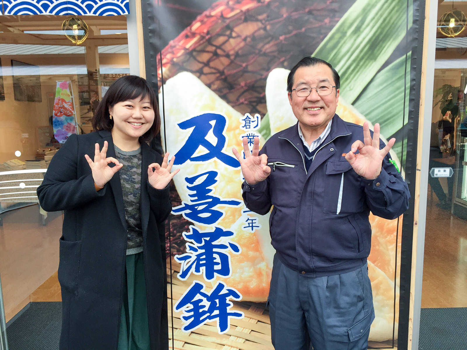 店舗の入り口横に下げられた「及善蒲鉾」と書かれた大きな布製の看板の前で、親指と人差し指で丸を作ったポーズをした及川さんと女性リポーターが並んで立っている写真