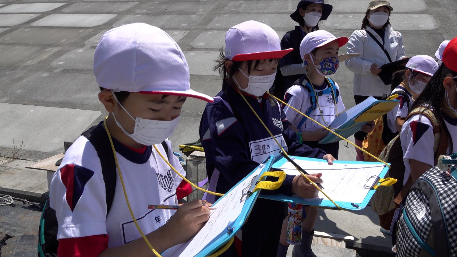 屋外で赤白帽子を被り体操服姿の児童たちが、男性に質問した内容などをクリップボードにメモを取っている様子を写した写真