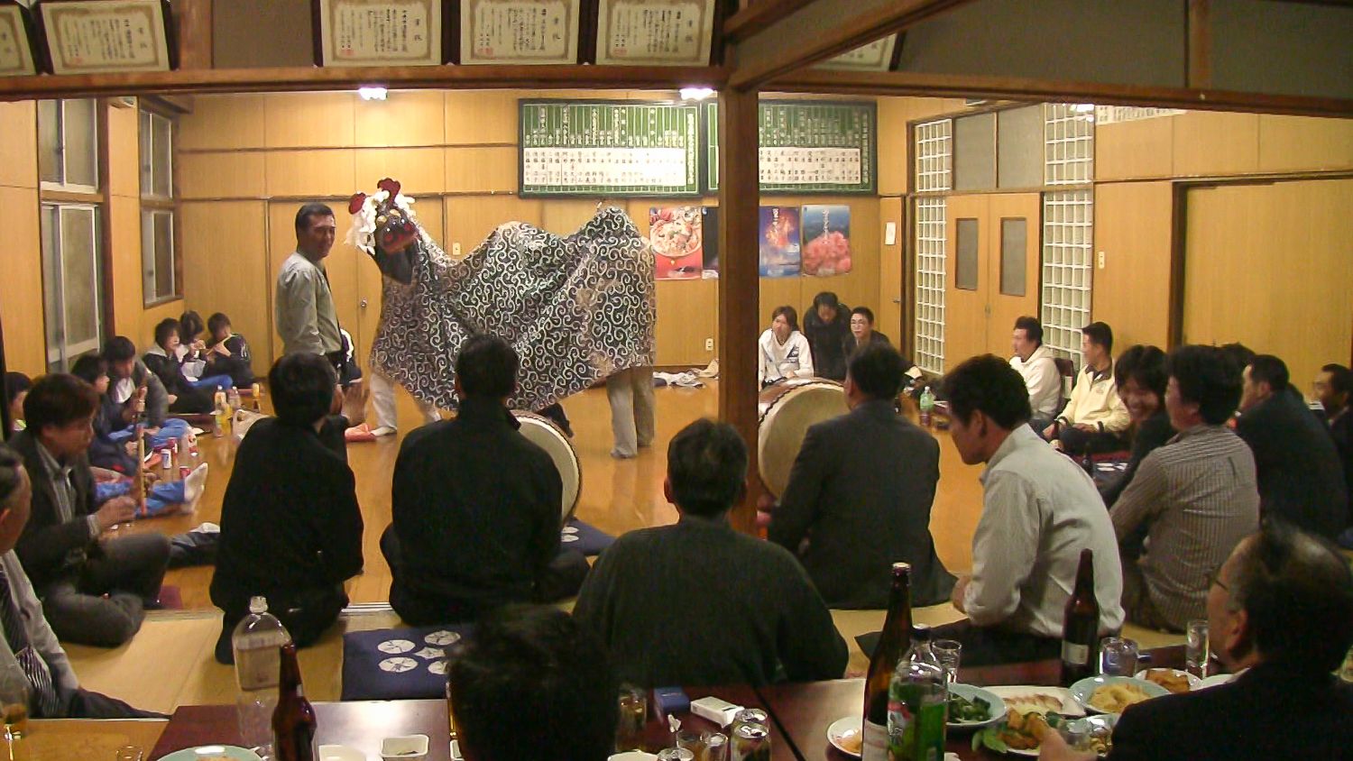 畳敷きの部屋で大勢の人々が囲む中、緑の模様の布を被った獅子舞が太鼓と笛の演奏に合わせて踊っている様子の写真