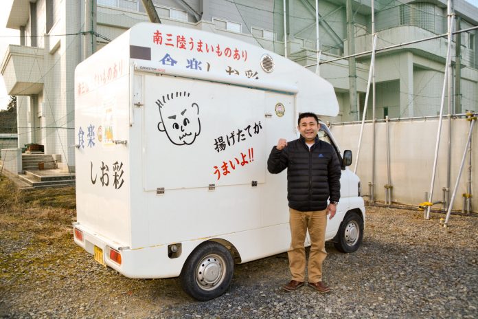 似顔絵や「揚げたてがうまいよ！！」の文字がかかれた白い移動販売車の横で黒いジャンパー姿の後藤さんが拳を作った右手を肩の高さまで挙げ笑顔で立っている写真