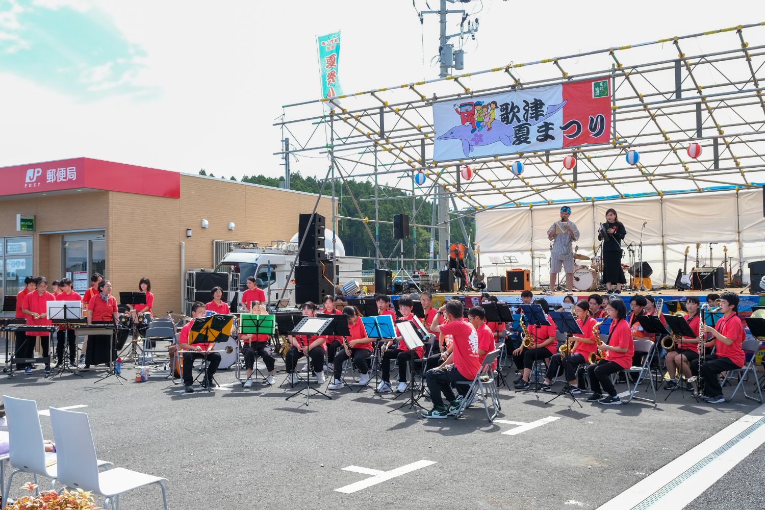 野外の駐車場で赤いポロシャツを着た多くの吹奏楽団員が椅子に座り演奏準備をしていて、背景に「歌津夏まつり」の横断幕があるステージが見える写真