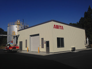 「AMITA」と赤い文字で書かれた白い外壁の四角い建物の後ろに銀色の大きな円柱のタンクが見える南三陸バイオガス施設の写真
