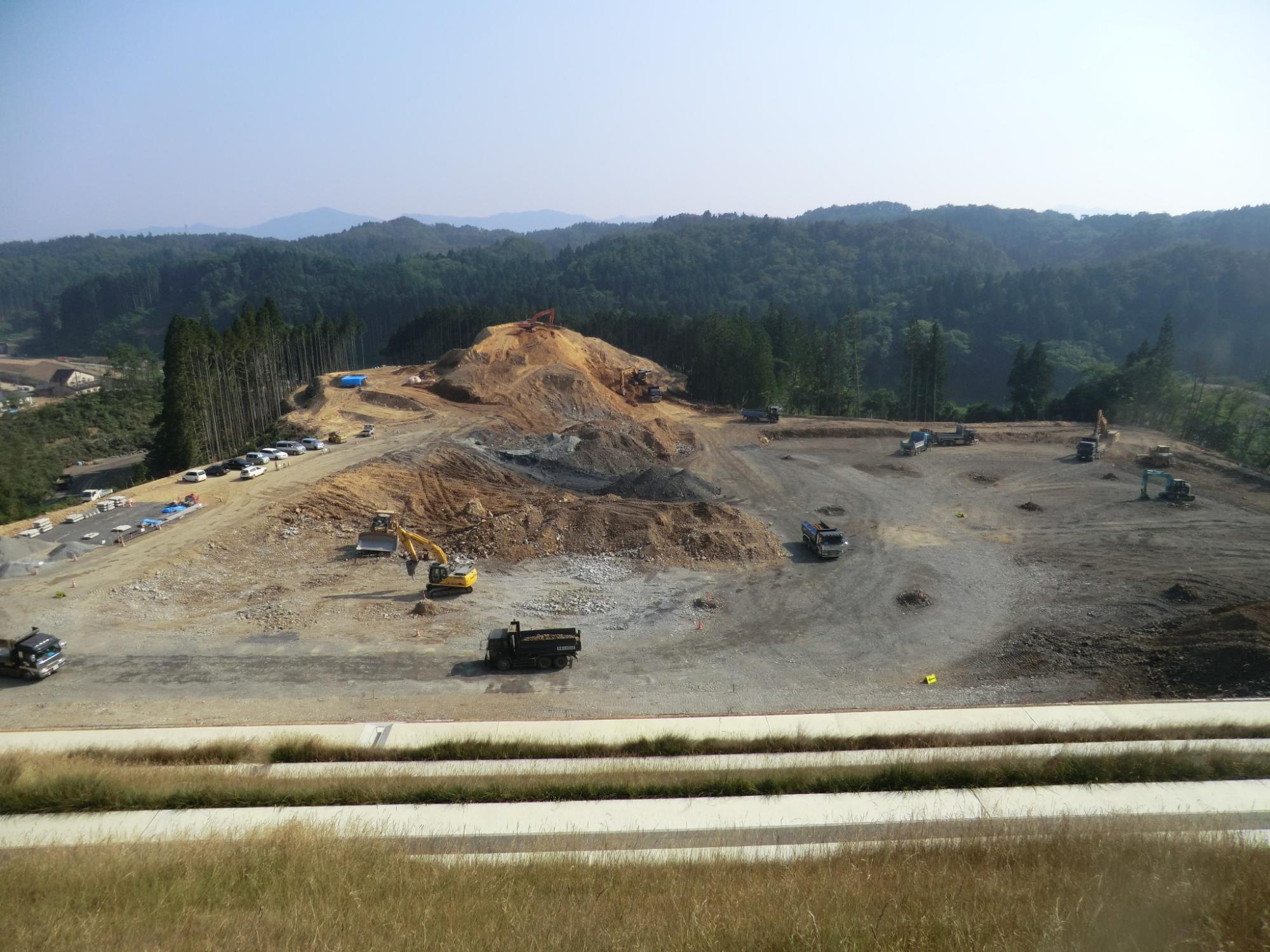 奥の土の山が少しずつ崩され整地がさらに進み、中央から右側の敷地の外側に窪みができ重機の作業範囲が広がった山中の工事現場の写真