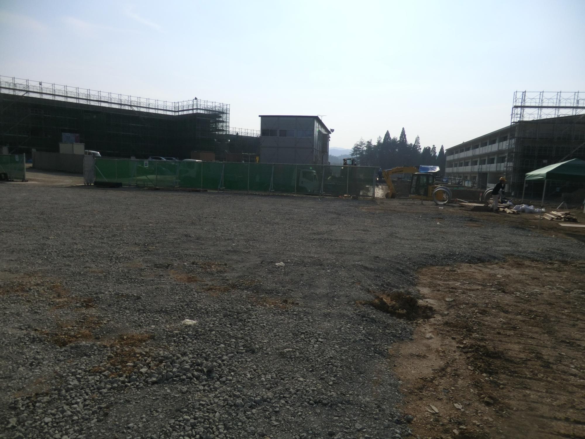 緑色の仮囲いの内側でショベルカーが動き、仮設建物と奥に立ち並ぶ建物が写った建設現場の写真