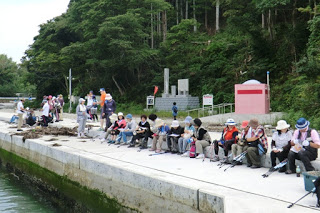 たくさんの参加者たちが、森や石碑を背景に、海に面したブロックに座り休憩している写真