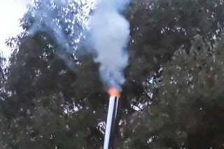 木の背景を背に、煙突の先端から白煙と共にオレンジ色の炎が噴き出している様子を写した、炭焼き工程でのガス燃焼の写真