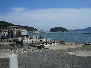 海に面したがれきの残るコンクリート基礎の跡地が広がり、左奥には倒壊した建物、正面には小さな島と港が見える、海辺の様子の写真