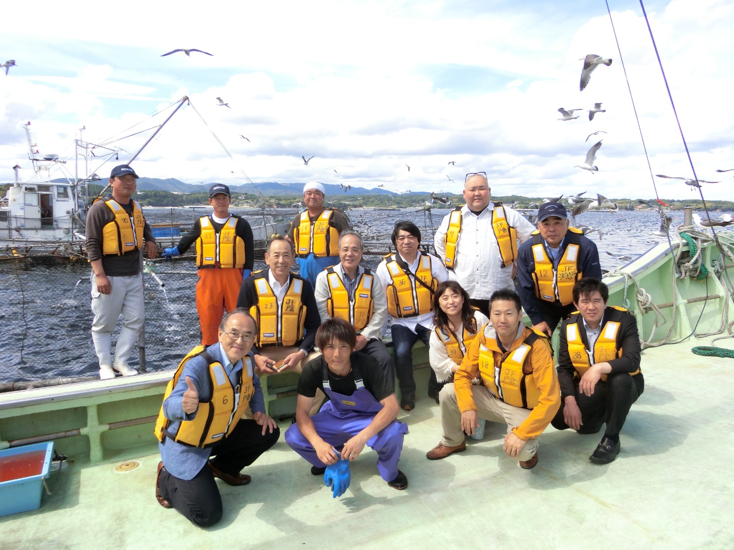 海に浮かぶ養殖いかだの前で、救命胴衣を着た「Fish & Dish Project」のメンバーたちが船の上で集合写真を撮っている写真