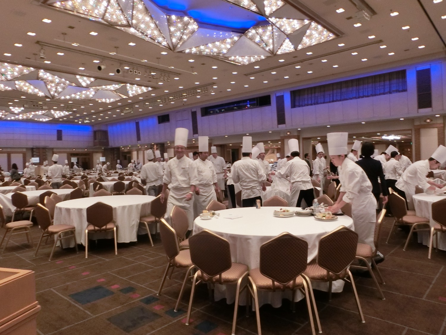大きな宴会場で、白い帽子にコックコートを着た多くのシェフが丸テーブルの間を行き来しながら料理の準備を進めている写真