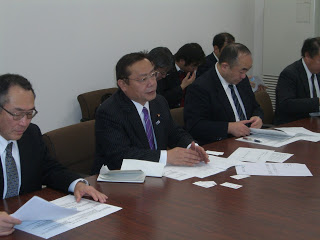 会議室の中でスーツを着た数人の関係者の方々が資料が広げられた長テーブルに座り、谷副大臣が話をしている様子の写真