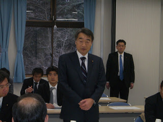 会議室の中で、関係者の方々が席についており、中央に立っている根本大臣が話をしている様子の写真