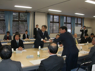 スーツ姿の複数の関係者の方々が座って見守る中、佐藤町長が根本復興大臣に要望書を手渡ししている様子の写真