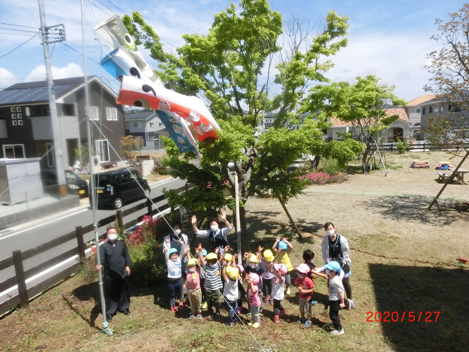 2020年5月27日に撮影され、木々や花が咲いている広々とした園庭で、子どもたちと保育士がこいのぼりの下に立ち、カメラに向かって手を振っている様子の写真