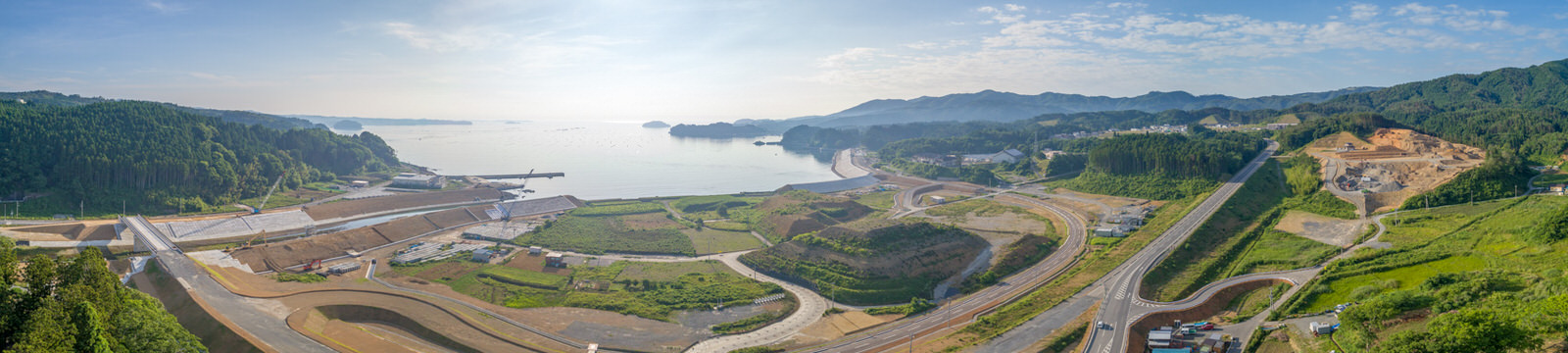 緑の丘と森林に囲まれた海沿いの地域を広く見渡した風景で、中央奥に水面が広がり、手前には道路や建設現場、建物が見え、空には雲が少し浮かぶ晴れた日のパノラマ写真
