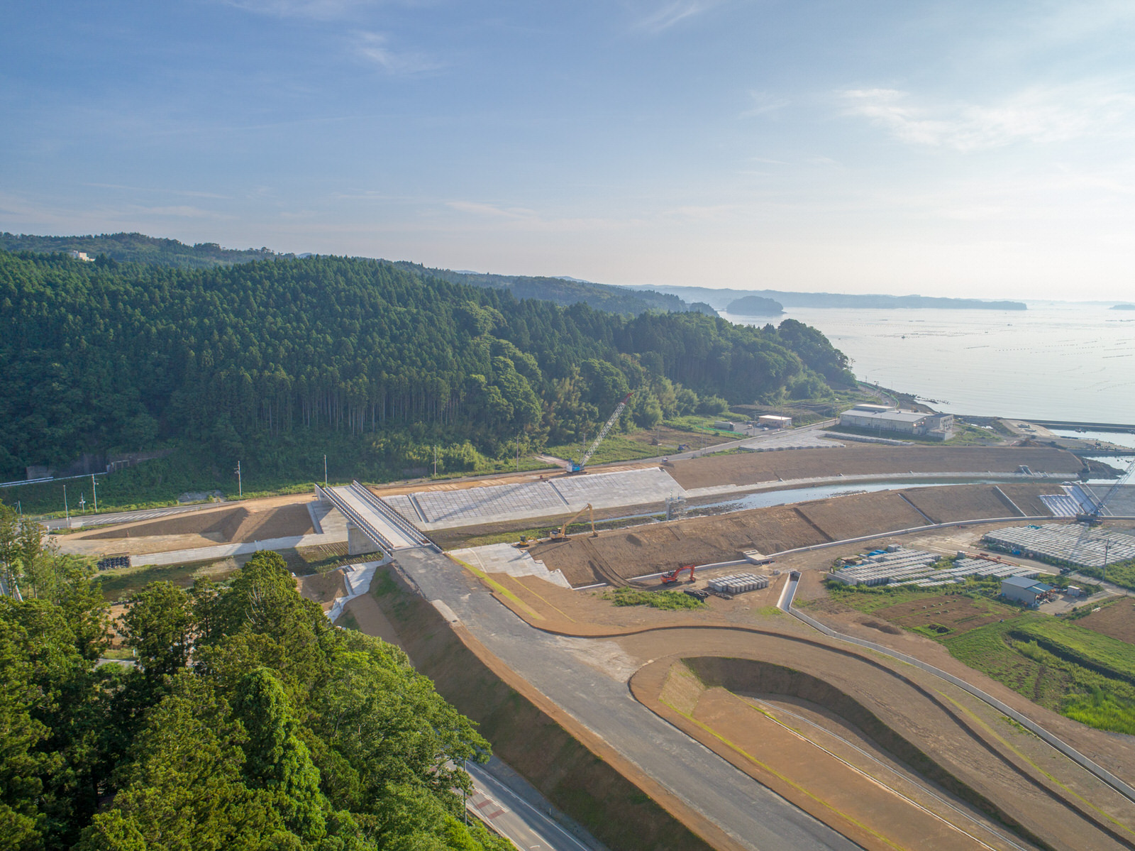 海に面した地域で、新しく建設された、または建設中のインフラ施設があり、道路や橋、防災や埋め立てに関する構造物が見られ、背景には森林に覆われた丘と水平線まで広がる海が写っている写真