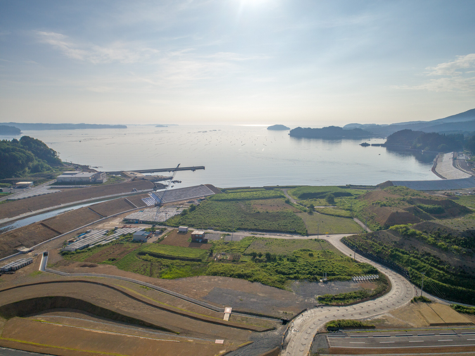 開発された土地と自然が混在する海沿いの地域を上空から撮影したもので、道路や緑の野原、海岸近くの建設地が見え、穏やかな海には小さな島々が点在し、太陽の柔らかな光が風景を照らしている写真