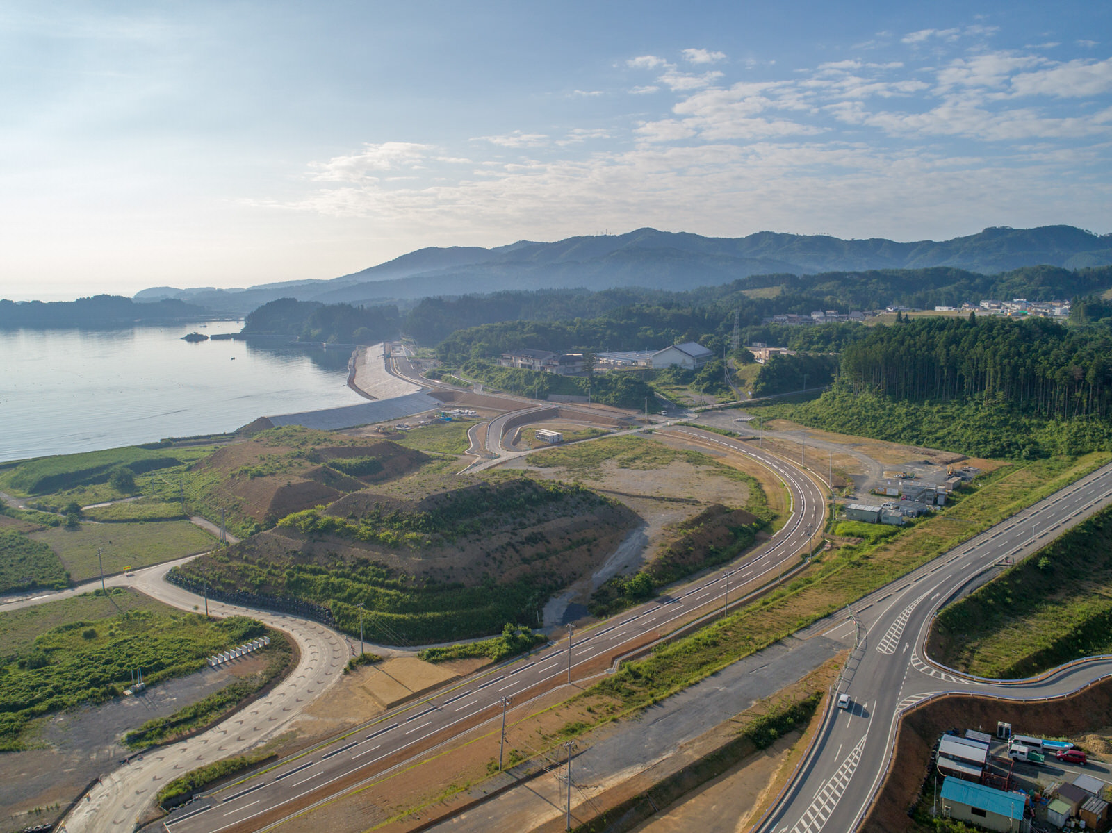 緑の丘と森林に囲まれた海沿いの地域を上空から撮影したもので、曲がりくねった道路が地形に沿って走り、遠くには建物や構造物が点在し、背景には山々と雲が浮かぶ空が広がる写真