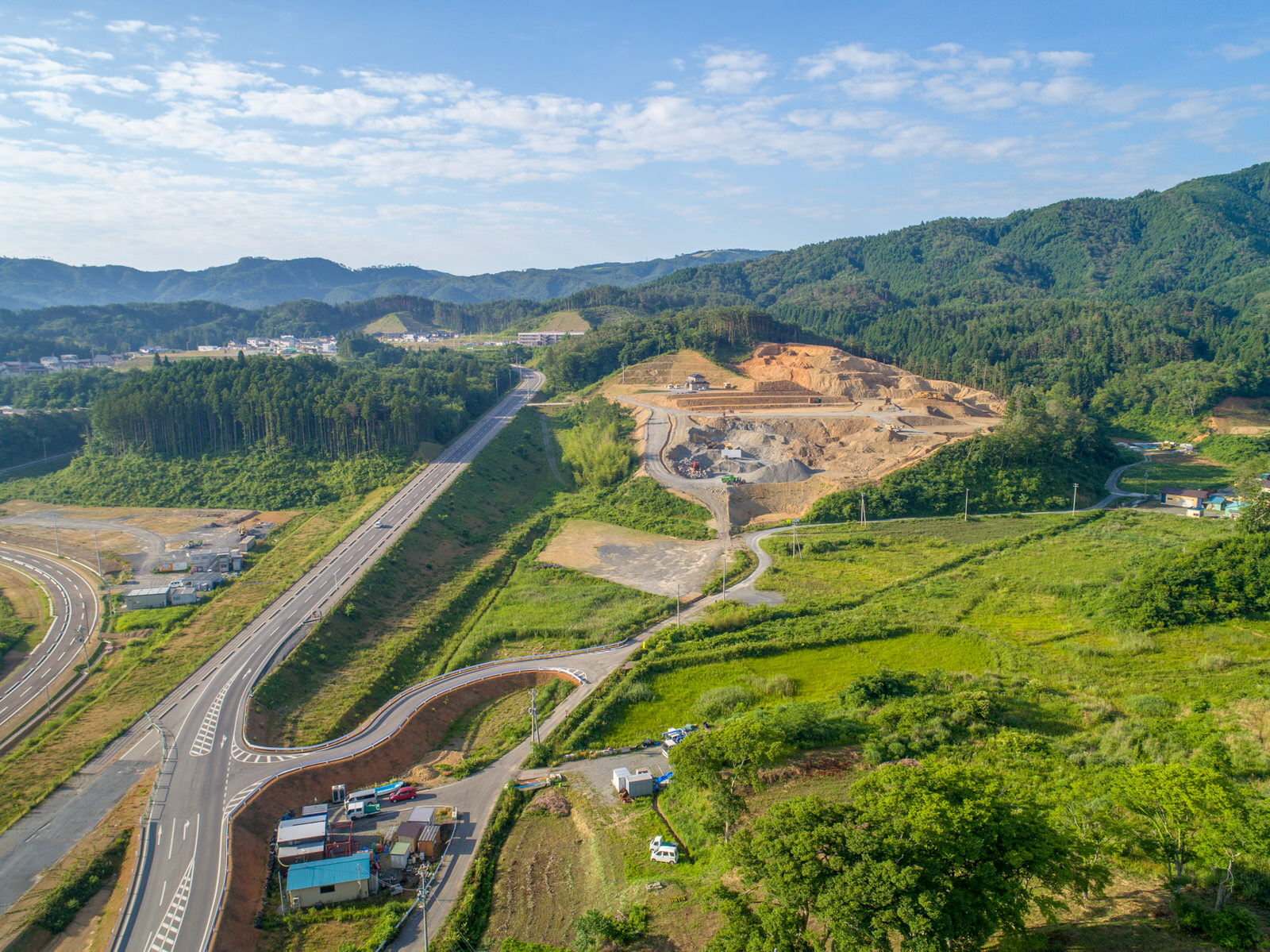 緑豊かな丘の上にある建設現場を写した風景で、背景には山々が広がり、道路が敷地を通り抜け、画像の下部には複数の車両や建物が見え、空には点在する雲が浮かぶ晴れた日の工事現場を撮影した写真