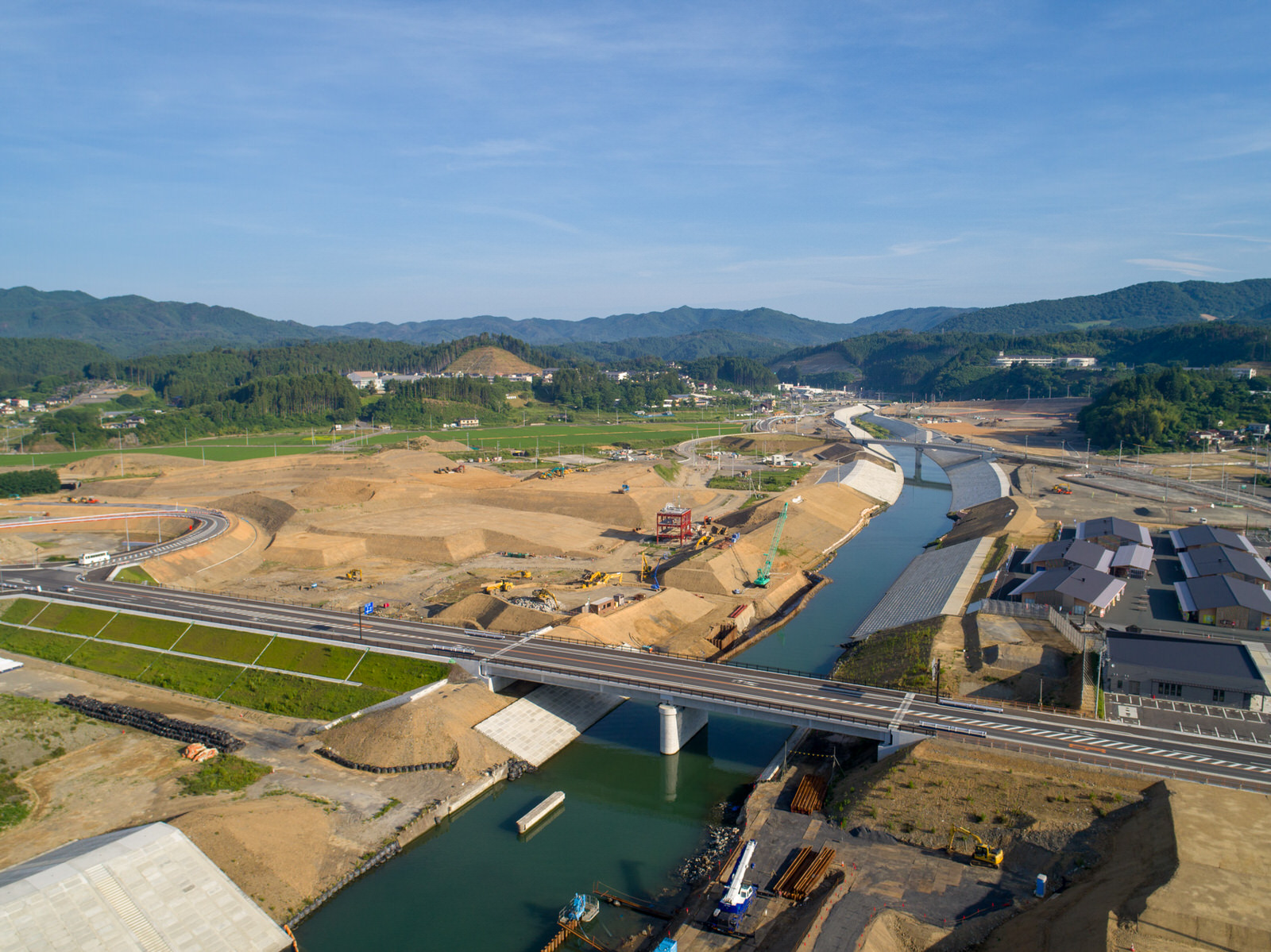 川が流れる左側の建設現場を上空から撮影したもので、川には橋が架かり、両岸には建設機材や資材が並び、周囲には建物や広い野原、背景には丘や山が広がる建設現場の写真