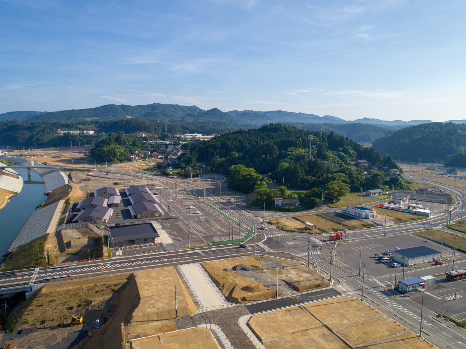 自然と開発が混在する志津川地区の風景を上空から撮影したもので、手前には複数の建物や道路、駐車場があり、左側には川が流れ橋が架かっており、背景には森林に覆われた丘と山々が澄んだ青空の下に広がる志津川地区の写真