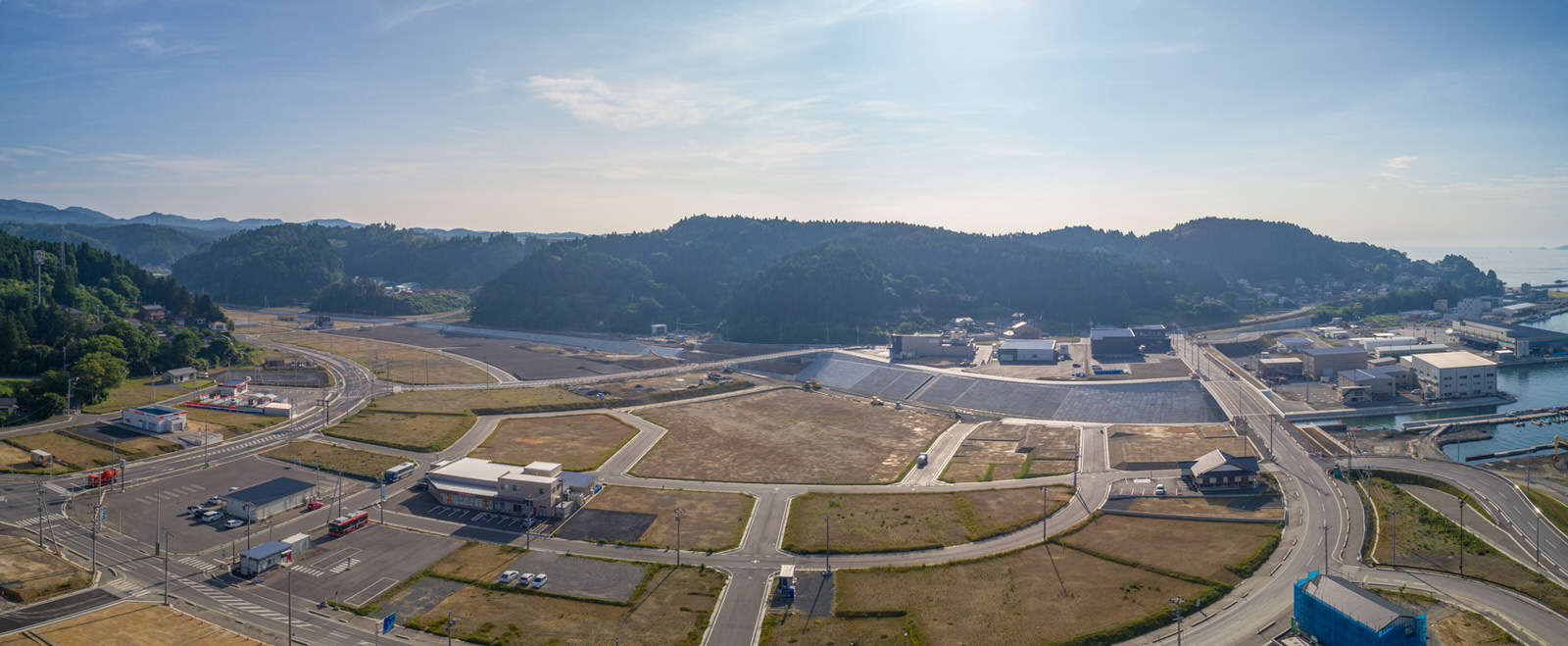 開発された地域と自然が混在する町を上空から撮影したもので、複数の建物や道路、空き地が見られ、町は丘と森林に囲まれ、背景には海岸線が広がり、晴れた空に雲が少し浮かぶパノラマ写真