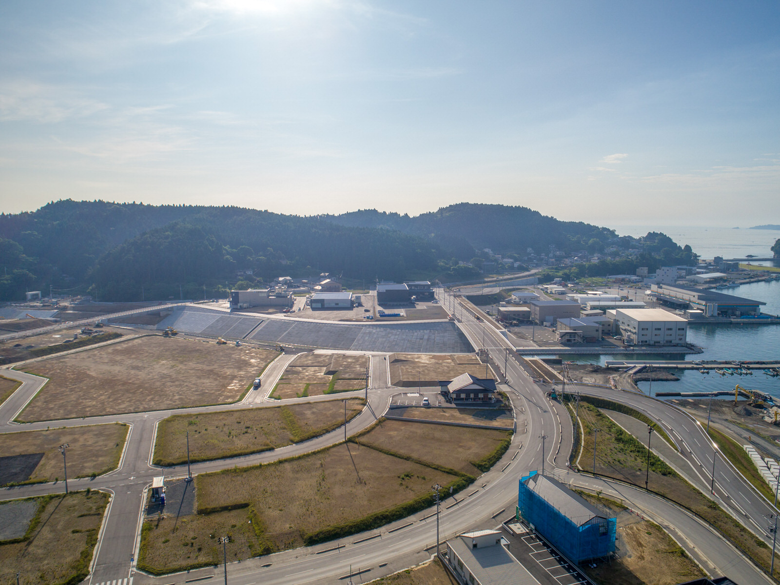 開発された土地と自然が混在する海沿いの地域を上空から撮影したもので、複数の建物や道路、空き地が点在し、右側には海岸線が広がり、水面が遠くまで続き、背景には森林に覆われた丘が澄んだ空の下に広がる写真