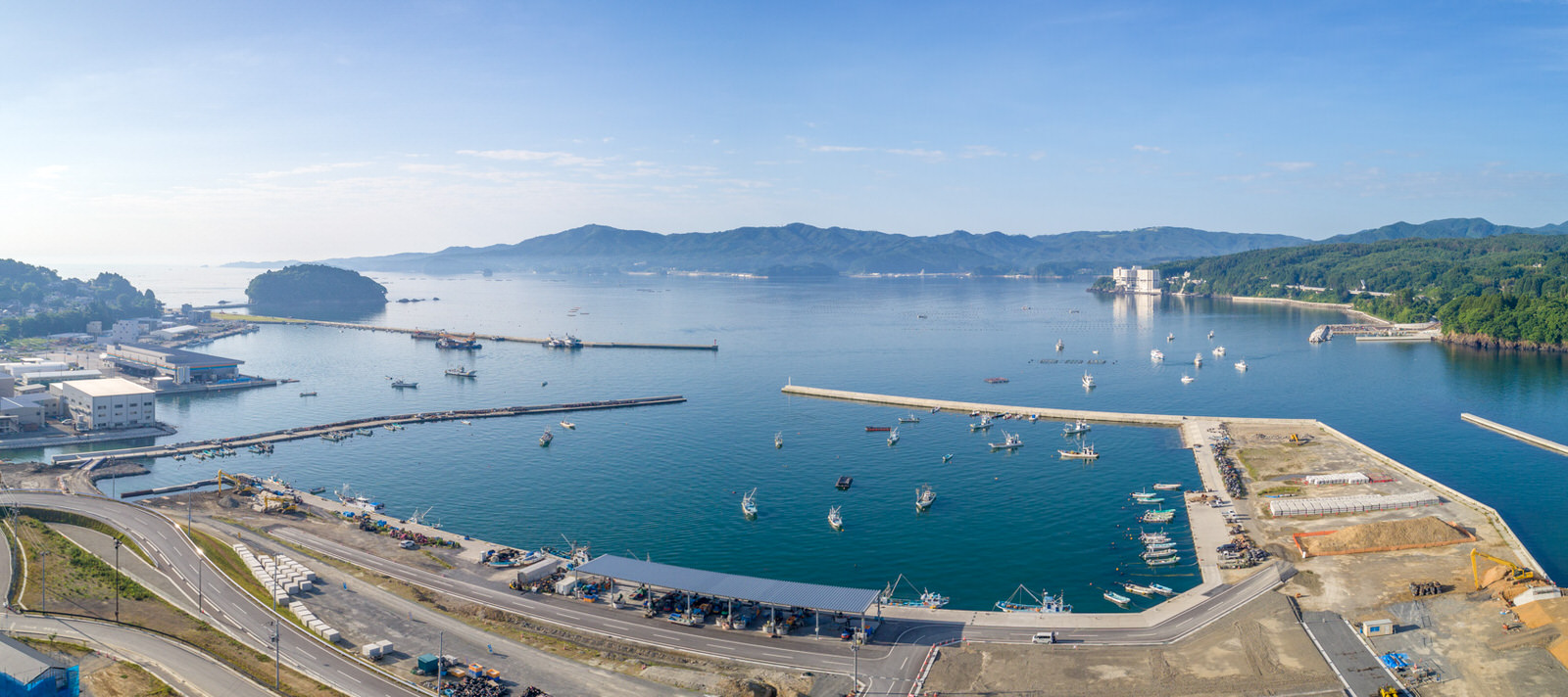 港のある海沿いの地域を広く見渡した風景で、水面には複数の船が停泊し、海岸沿いには建物やインフラが並び、背景には山々と澄んだ青空が広がるパノラマ写真
